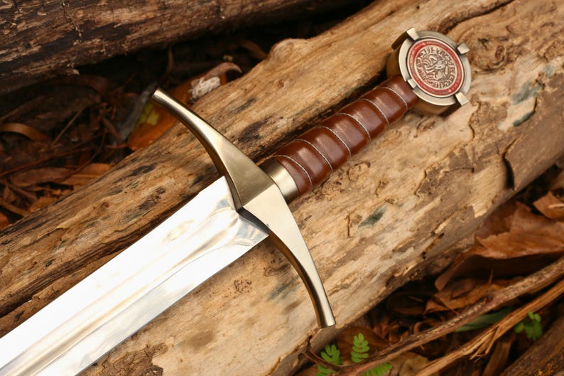 Puede incluir: Imagen detallada de una espada con una hoja plateada pulida y un mango envuelto en cuero marr&oacute;n. La guarda y la empu&ntilde;adura son de color dorado. El pomo presenta un dise&ntilde;o circular con detalles rojos y dorados. La espada descansa sobre troncos de madera.