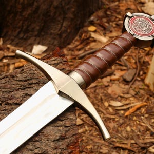 Puede incluir: Una espada de estilo medieval con una hoja de acero pulido y un mango envuelto en cuero marr&oacute;n. La empu&ntilde;adura presenta una guarda met&aacute;lica decorativa y un pomo con un emblema circular. La espada descansa sobre una superficie de madera envejecida.