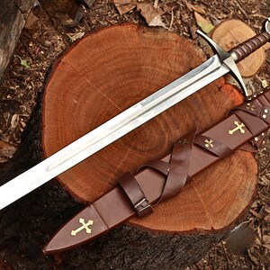 Puede incluir: Una espada de estilo medieval con una hoja plateada y un mango envuelto en cuero marr&oacute;n. La espada descansa sobre una superficie de madera junto a una vaina de cuero marr&oacute;n adornada con cruces doradas. La vaina tiene una correa.