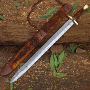 May include: A handcrafted sword with a patterned steel blade and a wooden handle. The sword lies next to its wooden sheath, which features intricate carved designs and leather accents. The overall colour scheme is brown and silver.