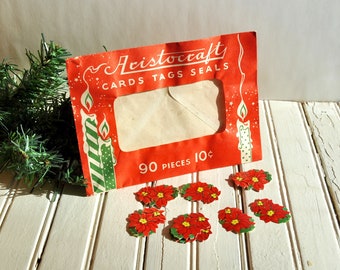 May include: Vintage red Aristocraft card package with the text "CARDS TAGS SEALS" and "90 PIECES". The package features illustrations of candles and is accompanied by several red poinsettia cutouts with green leaves. The items are on a white wooden surface.