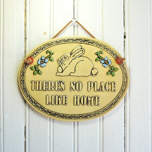 May include: A yellow oval ceramic sign with a white rabbit and flowers. The sign reads "There's no place like home".