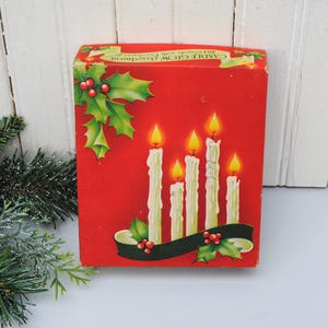 May include: A red box featuring a Christmas design with illustrations of lit candles, holly, and berries. The box has the words "CANDLE GLOW Christmas" printed on it. The box is rectangular and has a white wooden background.