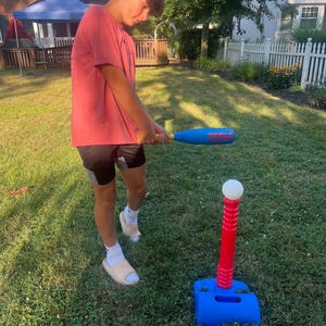 May include: A person holding a blue and yellow baseball bat, preparing to hit a baseball off a red and blue tee. The tee is on a green lawn. The bat has the word "CARLO" printed on it.