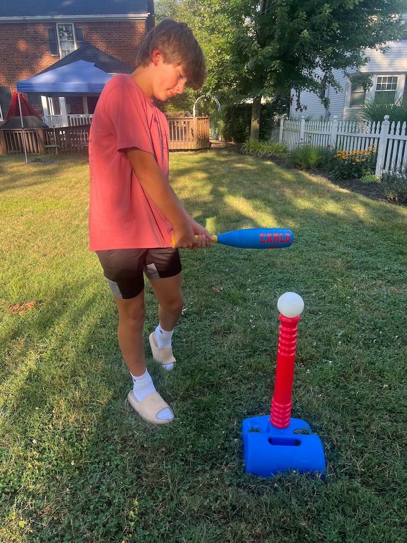 May include: A person holding a blue and yellow baseball bat, preparing to hit a baseball off a red and blue tee. The tee is on a green lawn. The bat has the word "CARLO" printed on it.