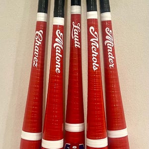 May include: A collection of five red and black baseball bats. Each bat has a black handle and a red barrel with a white stripe. The names Chavez, Malone, Lautt, Nichols, and Minder are printed on the barrels. The bats are labeled "Queen Creek Firefighters".