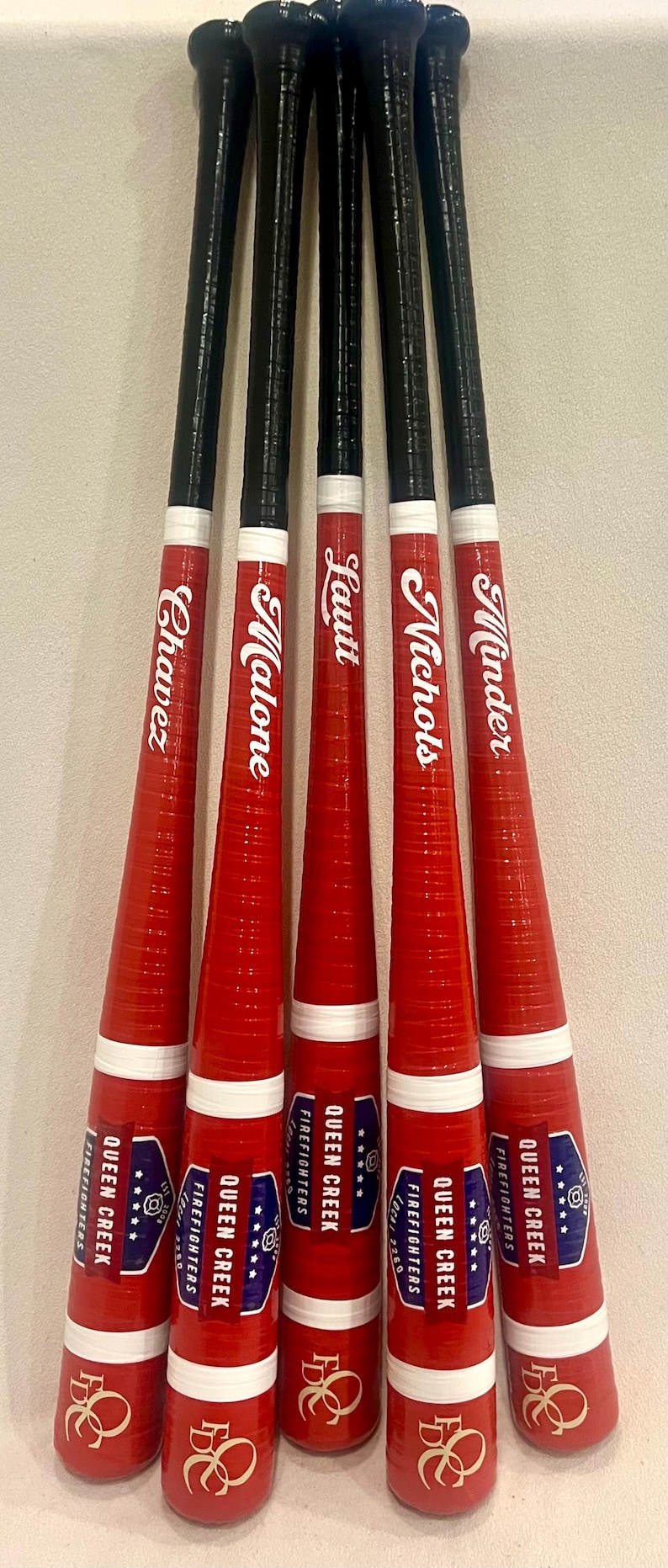May include: A collection of five red and black baseball bats. Each bat has a black handle and a red barrel with a white stripe. The names Chavez, Malone, Lautt, Nichols, and Minder are printed on the barrels. The bats are labeled "Queen Creek Firefighters".