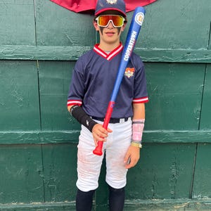 May include: A young baseball player in a navy blue uniform with red and white accents, holding a blue and red bat. The bat reads "Cooperstown". The player wears a matching cap, white pants, and protective eyewear. The background features a green wooden wall and a red, white, and blue banner.