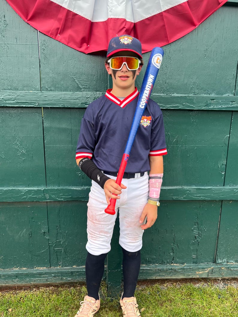 May include: A young baseball player in a navy blue uniform with red and white accents, holding a blue and red bat. The bat reads "Cooperstown". The player wears a matching cap, white pants, and protective eyewear. The background features a green wooden wall and a red, white, and blue banner.
