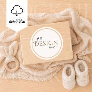 Puede incluir: Una caja de madera clara con un diseño circular "your DESIGN here" descansa sobre una manta beige. También hay un mordedor de madera con forma de conejito y patucos para bebé. La imagen incluye un icono de descarga digital.