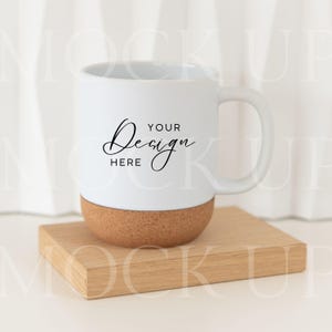 Könnte beinhalten: Weiße Keramiktasse mit Korkboden, die auf einem kleinen Holzquadrat steht. Die Tasse hat einen gebogenen Henkel und die Worte "YOUR Design HERE" in schwarzer Schrift. Die Tasse steht auf einer hellen Oberfläche.