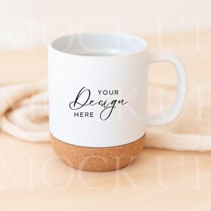 Könnte beinhalten: Weißer Keramikbecher mit Korkboden. Der Becher hat einen gebogenen Griff und die Worte "YOUR DESIGN HERE" in schwarzer Schrift. Der Becher steht auf einer hellen Holzoberfläche.