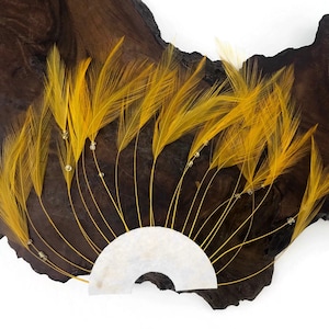 May include: A decorative hair accessory featuring a fan-shaped design. The piece is composed of vibrant yellow feathers, accented with small, clear beads. The feathers are attached to a white, semi-circular base, creating a striking visual contrast against the dark wood background.
