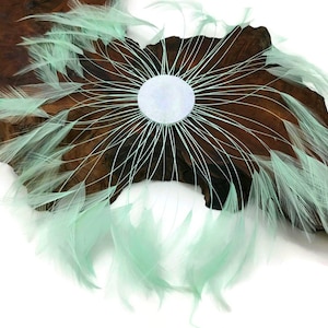 May include: A circular piece of art made with light green feathers and white string. The feathers are arranged in a radial pattern around a white circle in the center. The art is mounted on a piece of wood.