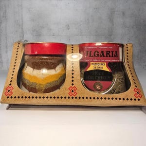 May include: A spice set featuring two glass jars with red lids, filled with colorful spices. The jars are presented in a decorative cardboard holder with a traditional design. One jar is labeled "Spice for Chiken".