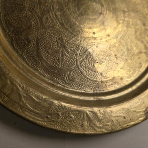 May include: A close-up of a gold-colored metal plate with intricate embossed floral and swirl patterns. The plate has a slightly raised rim and a circular design in the center. The surface has a worn, antique appearance.