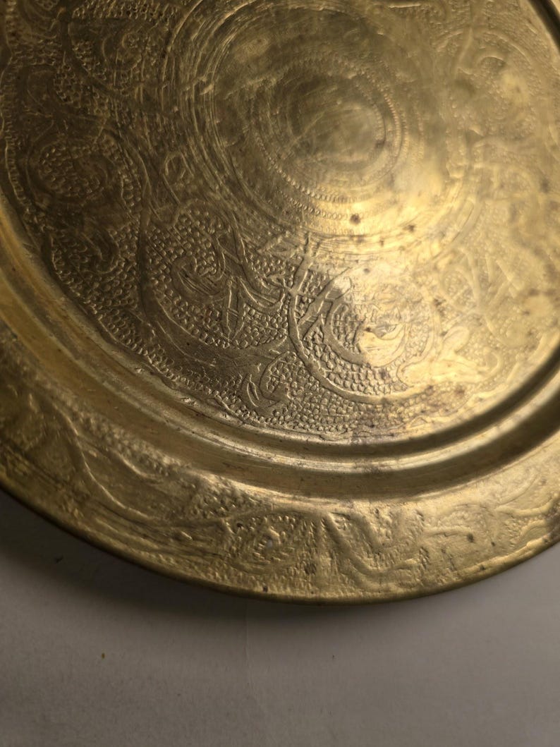 May include: A close-up of a gold-colored metal plate with intricate embossed floral and swirl patterns. The plate has a slightly raised rim and a circular design in the center. The surface has a worn, antique appearance.
