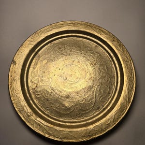 May include: A round, antique brass tray with an embossed floral design. The tray has a raised edge and a slightly worn appearance, suggesting age and use. The central design is a circular pattern with intricate details.