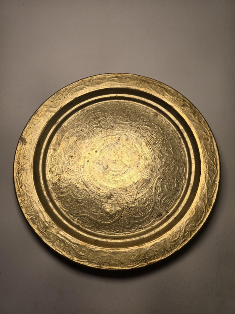 May include: A round, antique brass tray with an embossed floral design. The tray has a raised edge and a slightly worn appearance, suggesting age and use. The central design is a circular pattern with intricate details.