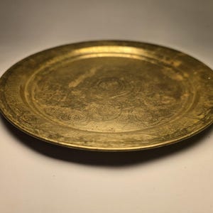 May include: A round, antique brass tray with an etched floral design. The tray has a slightly raised edge and a tarnished, aged appearance, suggesting it is vintage. The surface shows signs of wear, adding to its character.