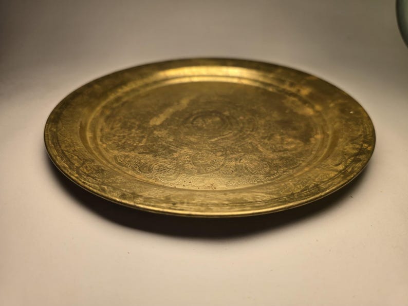 May include: A round, antique brass tray with an etched floral design. The tray has a slightly raised edge and a tarnished, aged appearance, suggesting it is vintage. The surface shows signs of wear, adding to its character.