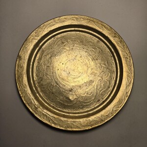 May include: A round, antique brass tray with intricate embossed floral and scrollwork patterns. The tray has a raised rim and a central design, showcasing a vintage aesthetic. The brass has a warm, golden hue.