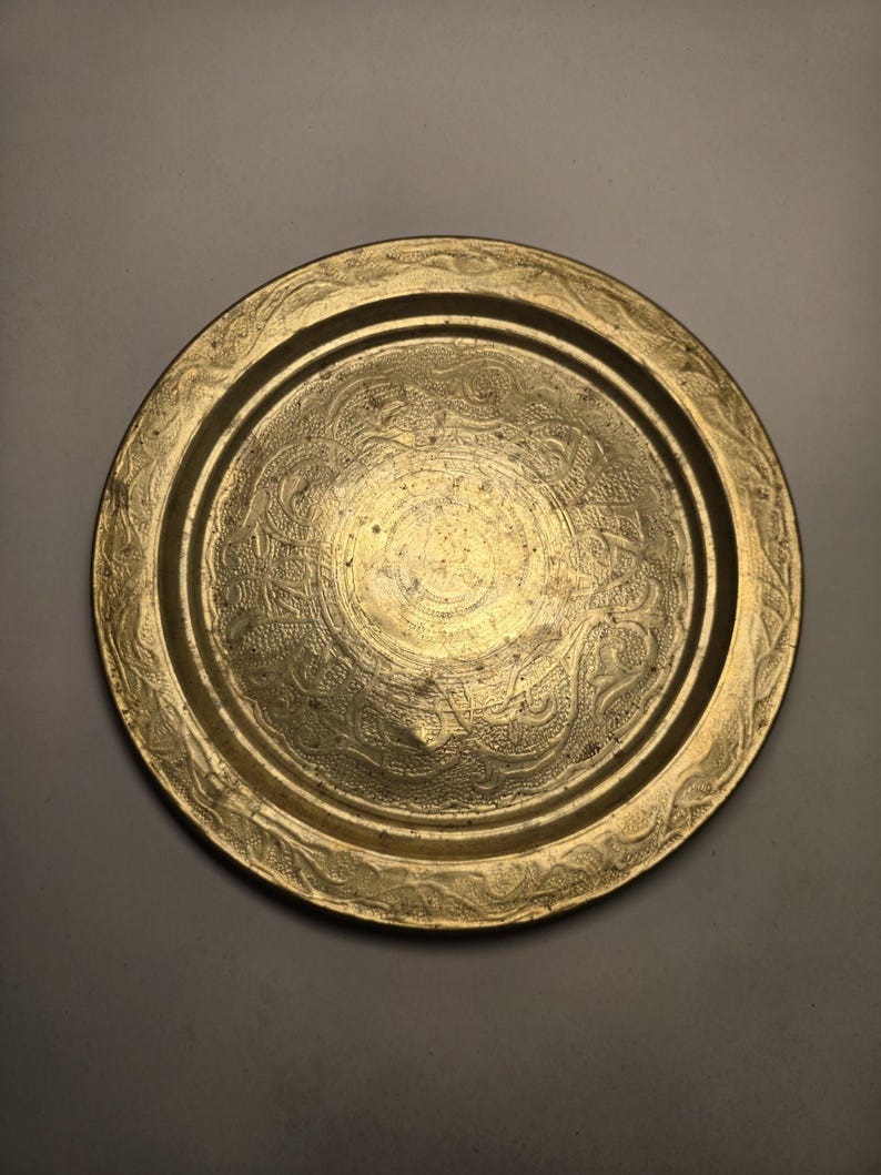 May include: A round, antique brass tray with intricate embossed floral and scrollwork patterns. The tray has a raised rim and a central design, showcasing a vintage aesthetic. The brass has a warm, golden hue.