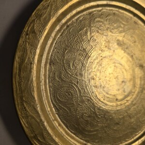 May include: A round, antique brass plate with intricate embossed floral and scrollwork patterns. The plate's surface has a warm, golden hue, with a slightly aged appearance. The design includes a detailed border and a central medallion.