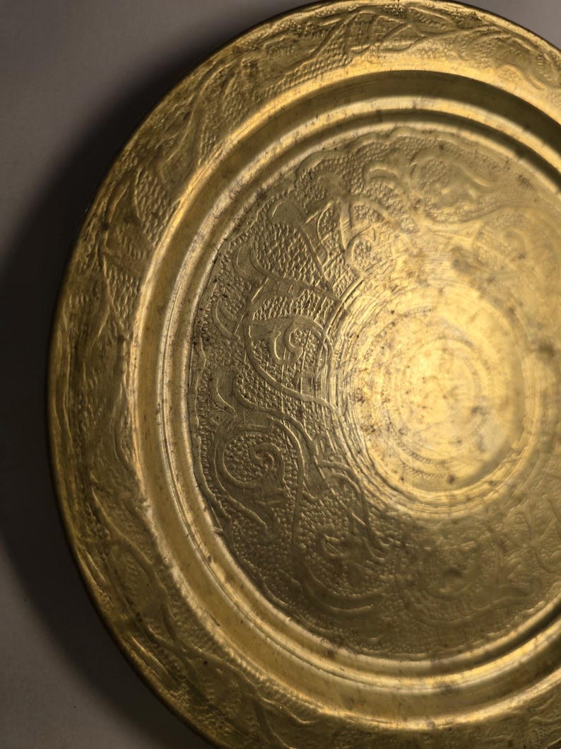 May include: A round, antique brass plate with intricate embossed floral and scrollwork patterns. The plate's surface has a warm, golden hue, with a slightly aged appearance. The design includes a detailed border and a central medallion.