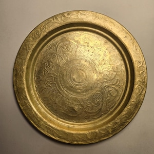 May include: A round, antique brass tray with intricate etched floral and scrollwork patterns. The tray has a raised rim and a central circular design. The brass has a warm, golden hue, and the surface shows signs of age.