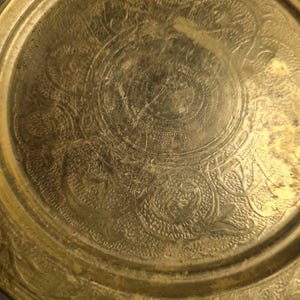 May include: A round, antique brass tray with intricate embossed floral and scrollwork patterns. The tray has a slightly worn, aged appearance, with a golden hue and visible surface imperfections. The design features a central medallion and a detailed border.