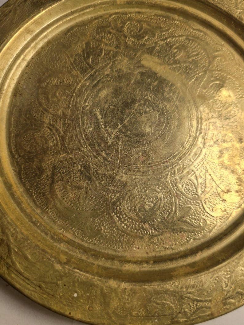 May include: A round, antique brass tray with intricate embossed floral and scrollwork patterns. The tray has a slightly worn, aged appearance, with a golden hue and visible surface imperfections. The design features a central medallion and a detailed border.