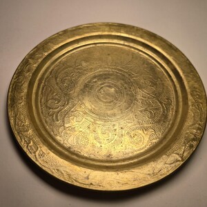 May include: A round, antique brass tray with intricate floral and scrollwork patterns. The tray has a raised edge and a central circular design. The brass has a warm, golden hue, and the details are finely etched.