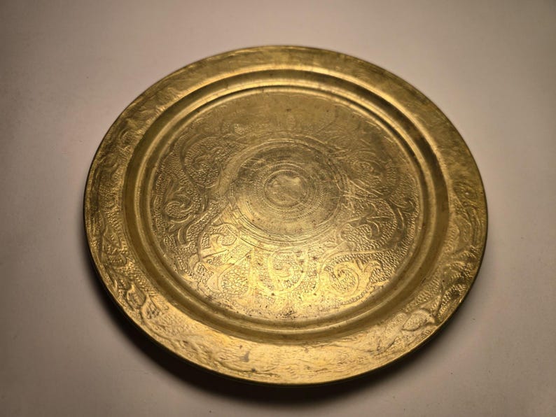 May include: A round, antique brass tray with intricate floral and scrollwork patterns. The tray has a raised edge and a central circular design. The brass has a warm, golden hue, and the details are finely etched.