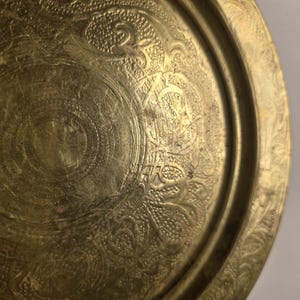 May include: A detailed close-up of a brass tray with intricate embossed floral and geometric patterns. The tray has a slightly tarnished, antique appearance, with a raised rim and detailed designs throughout the surface.
