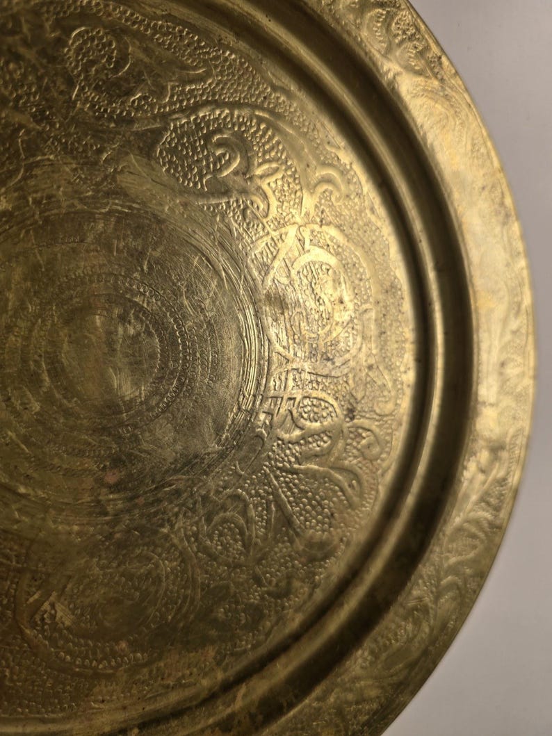 May include: A detailed close-up of a brass tray with intricate embossed floral and geometric patterns. The tray has a slightly tarnished, antique appearance, with a raised rim and detailed designs throughout the surface.