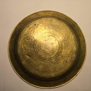 May include: A round, antique brass plate with an embossed floral design. The plate has a slightly raised rim and a golden hue, showing signs of age and use. The center features a detailed circular pattern.