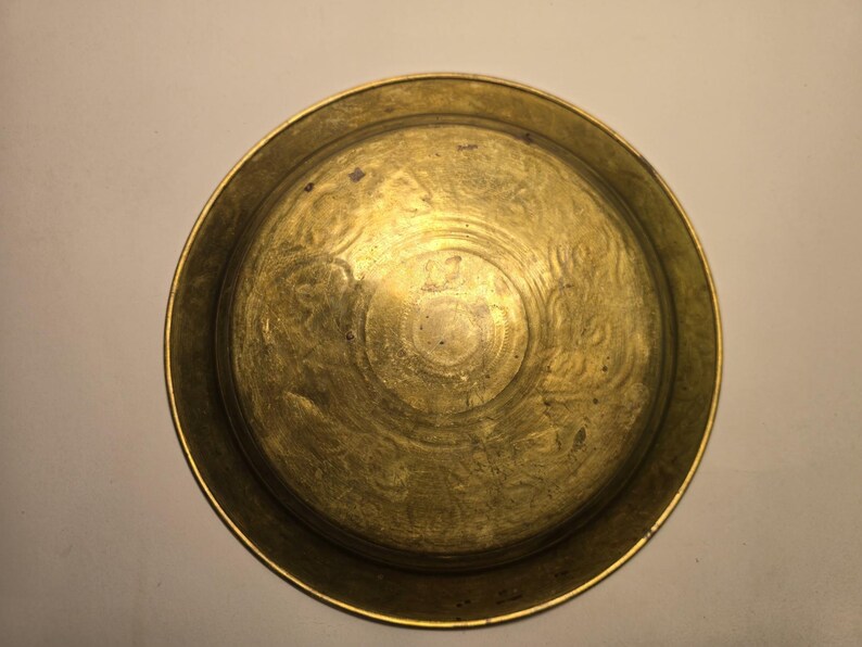 May include: A round, antique brass plate with an embossed floral design. The plate has a slightly raised rim and a golden hue, showing signs of age and use. The center features a detailed circular pattern.