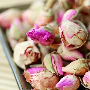 May include: A collection of dried rose buds featuring vibrant magenta and soft beige petals. The small, tightly closed flower heads are clustered together, displaying natural textures and varied shades of pink, cream, and muted green sepals.