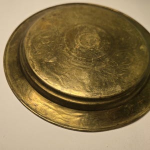 May include: A tarnished brass plate with a raised center and a wide, circular rim. The plate has a warm, golden hue and shows signs of age and wear, with visible scratches and imperfections across its surface. The design is simple and functional.