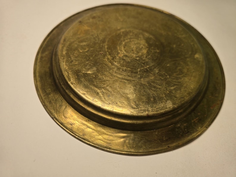 May include: A tarnished brass plate with a raised center and a wide, circular rim. The plate has a warm, golden hue and shows signs of age and wear, with visible scratches and imperfections across its surface. The design is simple and functional.