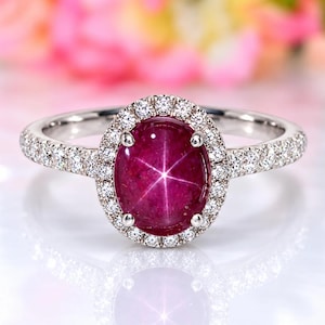 May include: A silver-toned ring featuring a central oval-shaped deep pink star ruby cabochon. The gemstone is surrounded by a halo of small round white diamonds, with additional diamonds set along the band on both sides of the centre stone.