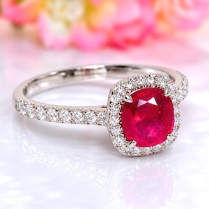 Puede incluir: Un anillo de tono plateado con una piedra preciosa roja central de corte cojín rodeada por un halo de pequeñas piedras blancas de corte redondo. La banda también tiene una hilera de pequeñas piedras blancas redondas en los lados.
