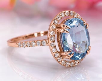 May include: A rose gold ring featuring a large, oval-cut light blue gemstone at the center. The stone is surrounded by a halo of small, round, clear white stones, with additional clear stones set along the band of the ring.