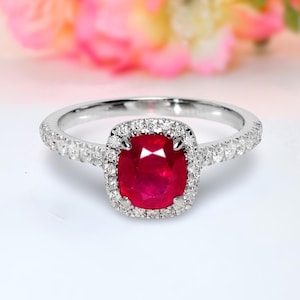 Pode incluir: Um anel de prata com uma grande pedra central de rubi com corte almofada, rodeada por um halo de diamantes menores. A banda também é cravejada com pequenos diamantes. Fundo desfocado rosa e branco.