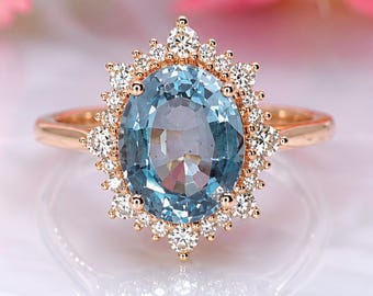 May include: A rose gold ring featuring a large oval-cut light blue center stone surrounded by a halo of small round white diamonds. The band is thin and polished, showcasing a classic prong setting that secures the central gemstone.