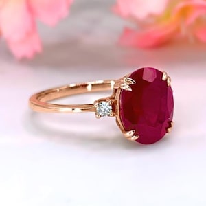 May include: Rose gold ring featuring a large, oval-cut ruby gemstone. The ring has a smaller, round diamond accent on one side. The ruby is a deep red color, and the ring is set against a soft, blurred background.