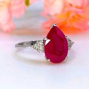 May include: A silver ring with a large, pear-cut ruby. The ruby is a deep red hue, with smaller, clear gemstones on either side. The ring is set against a soft, blurred background of pink and orange.