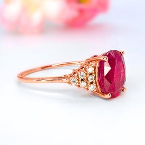 Puede incluir: Anillo de oro rosa con una gran gema de rubí de corte ovalado. El anillo está adornado con pequeños diamantes redondos en la banda. El rubí es de color rojo intenso y el anillo está sobre un fondo suave y borroso.