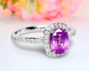 May include: A silver ring with a large, square-cut, vibrant purple gemstone at the center, surrounded by a halo of smaller, clear stones. The band is also adorned with small, clear stones. The background features soft pink and white flowers.